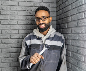 A happy, black male car mechanic with a beard, wearing work clothes and holding a wrench in his...