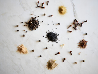 Overhead shot of various culinary spices and ingredients scattered artistically across a smooth white marble countertop. Includes black spices like pepper, black sesame, and clove. Emphasis on natural