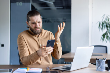 A frustrated man holds a smartphone, seemingly confused or exasperated while sitting at a desk. A...