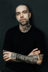 Tattooed man poses with crossed arms against a dark background, showcasing detailed arm tattoos and...