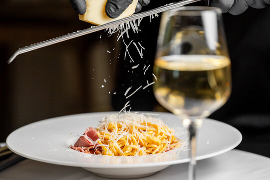 A chef in black gloves grates fresh cheese over a beautifully plated pasta dish while a glass of white wine adds elegance to the dining experience.