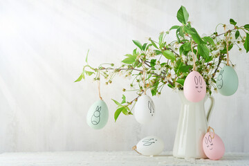Easter bouquet of cherry blossom branches decorated with funny eggs with drawings of hares and smiles in vintage vase against background of old concrete gray wall. Easter card. Copy space.
