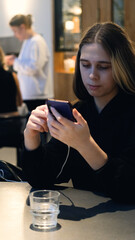 Young teenage girl sitting in modern cafe, using her smartphone to browse internet, check social media or send messages, enjoying the moment of digital connection in a cozy environment.