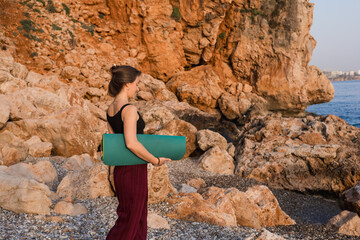 Young caucasian woman holding rolled up green yoga mat against background of sea rocks.