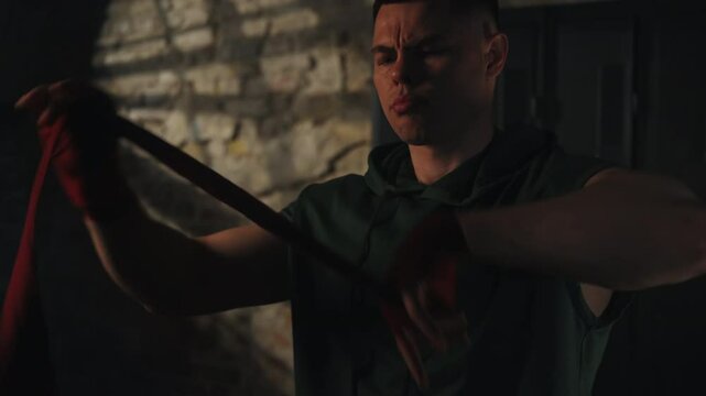 Man in sleeveless hoodie and red hand wraps tightens bandages on fists in dimly lit gym with brick walls, preparing for intense workout. Concept: discipline, preparation