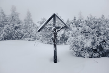 A dream like picture of frozen wooden cross of Jesus Christ hidden deep in the snow covered nature evokes solitude, redemption and silence of the winter foggy forest in freezing weather in Czechia