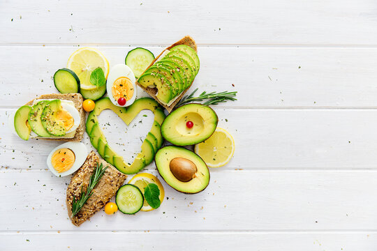 Overhead view of Avocado slices in a heart shape with brown bread, lemon and eggs on a wooden table