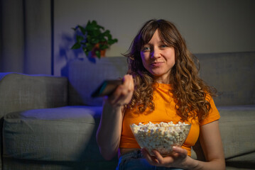 Young woman with curly hair is sitting on the floor in front of her sofa, holding a bowl of popcorn...