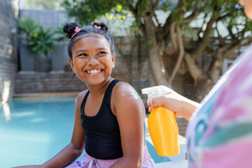 Happy woman applying sunscreen on smiling child at poolside, holding orange spray bottle