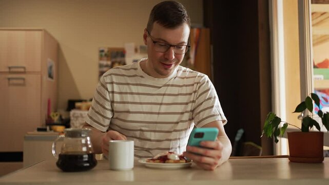 Young man enjoying morning routine, sitting at kitchen table while eating pancakes and sipping coffee, simultaneously browsing smartphone with relaxed demeanor