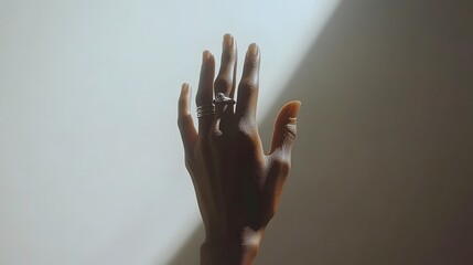 A close-up shot of a dark-skinned hand with rings, reaching up against a white background. Sunlight casts a dramatic shadow.