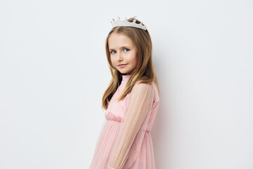 Young girl in a pink dress with a crown, looking confidently at the camera, set against a light...