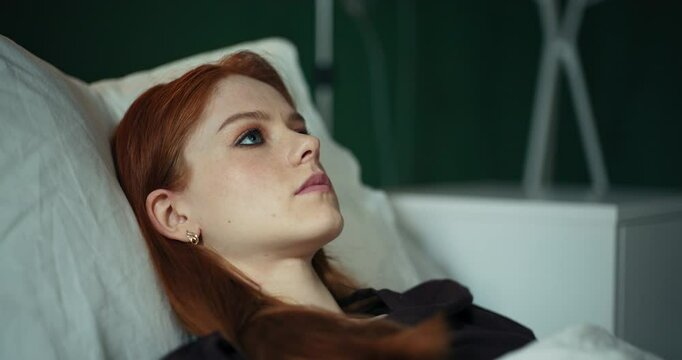 Young red-haired woman uses a meditation app on her phone while lying in a hospital bed, focusing on relaxation and mental well-being.