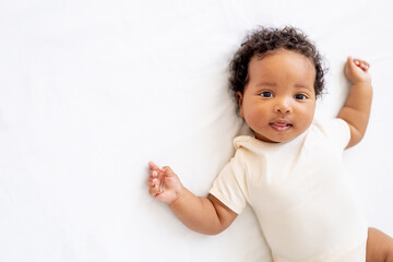 close-up portrait of a small African American baby girl in a white bodysuit on a cotton bed at...