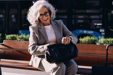 Elegant senior businesswoman in gray suit sits on bench, opening stylish black handbag. Female...