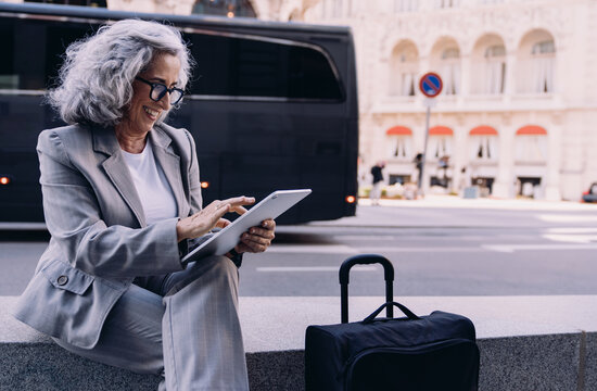 Sophisticated senior businesswoman sits on concrete bench, holding a tablet while observing the urban scene. Female suitcase beside her represents global business, travel, and modern work flexibility