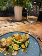 A beautifully arranged plate features crispy fried food paired with fresh cucumber slices and drizzled with sauce. A glass of white wine complements the meal, set on a wooden table