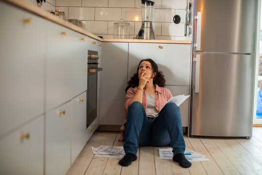 Stressed woman sitting on kitchen floor overwhelmed by bills