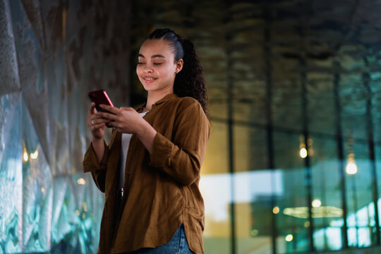 Confident Hispanic young woman scrolling through mobile phone, staying updated on social media and news. illuminated cityscape behind latin female emphasizes a connected, technology-driven lifestyle