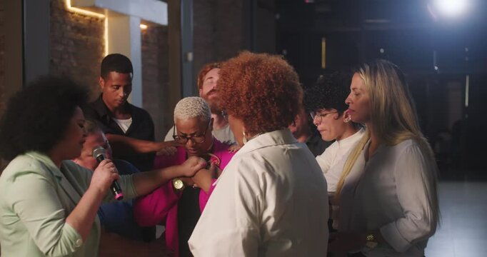Diverse group of people gathered in prayer around a Hispanic Latin woman of African descent in pink jacket, conveying unity, faith, and emotional connection in a spiritual setting