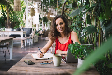 Woman in a red dress writing in a notebook at a tropical outdoor cafe, surrounded by greenery. A...