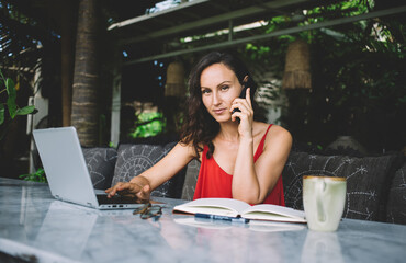 Focused freelancer in red dress multitasking on laptop and smartphone at a modern outdoor...