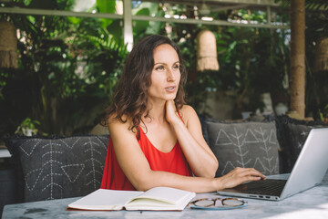 Focused woman working remotely on her laptop, sitting outdoors in a lush setting, wearing a red...