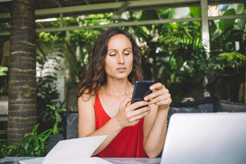 Focused woman in red top working remotely on her phone, with laptop and notebook nearby in a lush,...