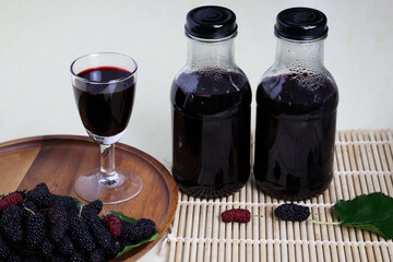 Two glass bottles and a long stem glass of dark red fruit juice are placed next to ripe mulberries...