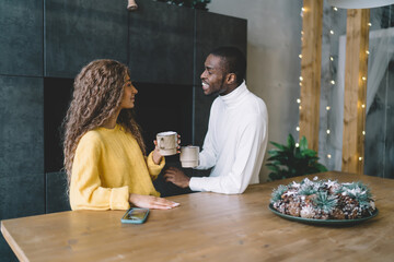 Happy African American couple enjoying coffee together in cozy, warmly lit home interior. woman listens attentively, while man in white turtleneck smiles, creating relaxed, intimate atmosphere