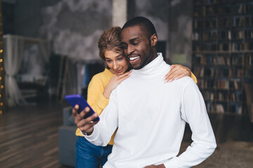 Smiling couple using an online banking app on a smartphone, embracing each other in a cozy, modern home. The dark-skinned man and his partner share a joyful moment while managing finances together
