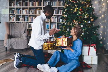 A joyful couple exchanges Christmas gifts by a beautifully decorated tree, surrounded by twinkling...