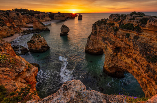 Dramatic cliffs and rock formations at sunset in Algarve, Portugal, with a vibrant sky and tranquil waters.