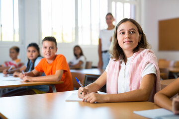 Young girl sitting at desk with her classmates during lesson in school and writing in copybook.