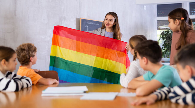 Young female teacher showing flag of LGBT to schoolchildren preteens and explaining LGBTQ social movements in classroom