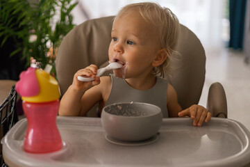 a blonde baby eats with a spoon while sitting at a children's dining table