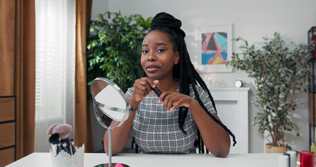 A makeup artist with dreadlocks paints her eyelashes while sitting at her desk. She records a vlog, sharing mascara benefits and beauty tips with her online audience.