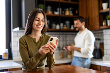 Happy woman using her smartphone while man cooking in the background, enjoying a moment together in a modern, stylish kitchen