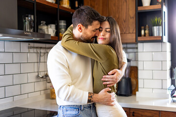 Romantic couple hugging in their modern kitchen, enjoying a quiet moment of affection and...