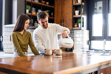 Happy couple preparing steaming hot drinks together in a modern kitchen, using an electric kettle...