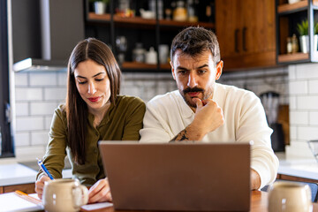 Young couple calculating domestic bills using laptop and taking notes, concerned about home finances