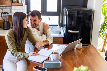 Young smiling couple working from home, using laptop and taking notes while sitting at kitchen table