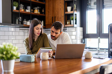 Young couple collaborating on a laptop and jotting down notes while working comfortably from their modern kitchen during the day