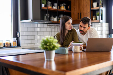 Young couple calculating domestic finances using laptop and notebook, managing their budget and expenses in their modern kitchen