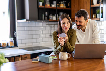 Young smiling couple is making online shopping using laptop and credit card while having breakfast in the kitchen