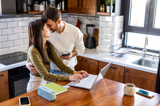 Young couple sharing a kiss while working together on a laptop in their cozy kitchen, enjoying a moment of love and connection - Powered by Adobe
