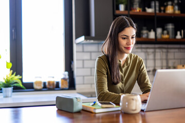 Young woman working remotely using laptop in her kitchen, enjoying a cup of coffee or tea