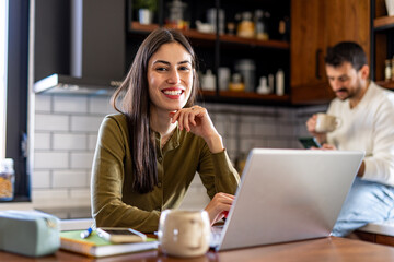 Smiling businesswoman using laptop, working from home, sitting at kitchen table, with husband in background drinking coffee