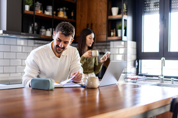 Man smiling and working from home, reviewing documents in his kitchen while a woman drinks coffee...