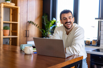 Cheerful businessman working remotely in a modern kitchen, enjoying productivity while using his...
